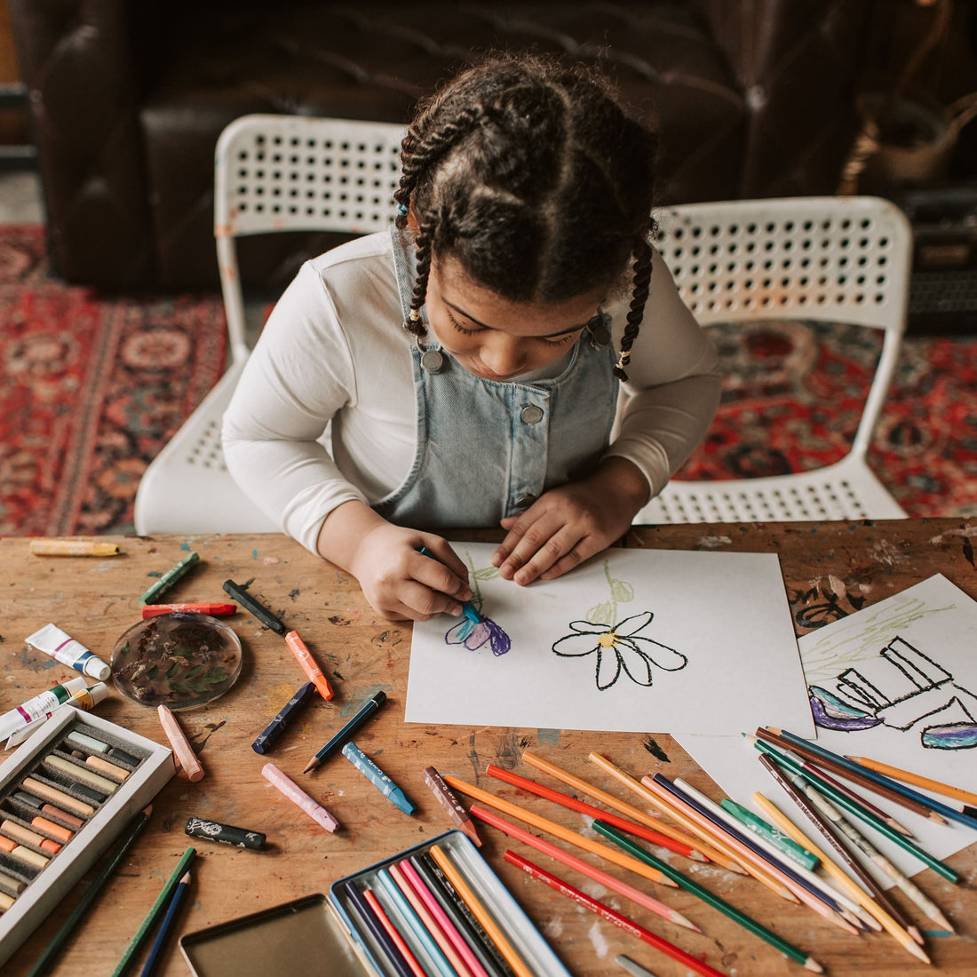 Menina sentada em uma mesa cheia de materiais de colorir, desenhando em uma folha.